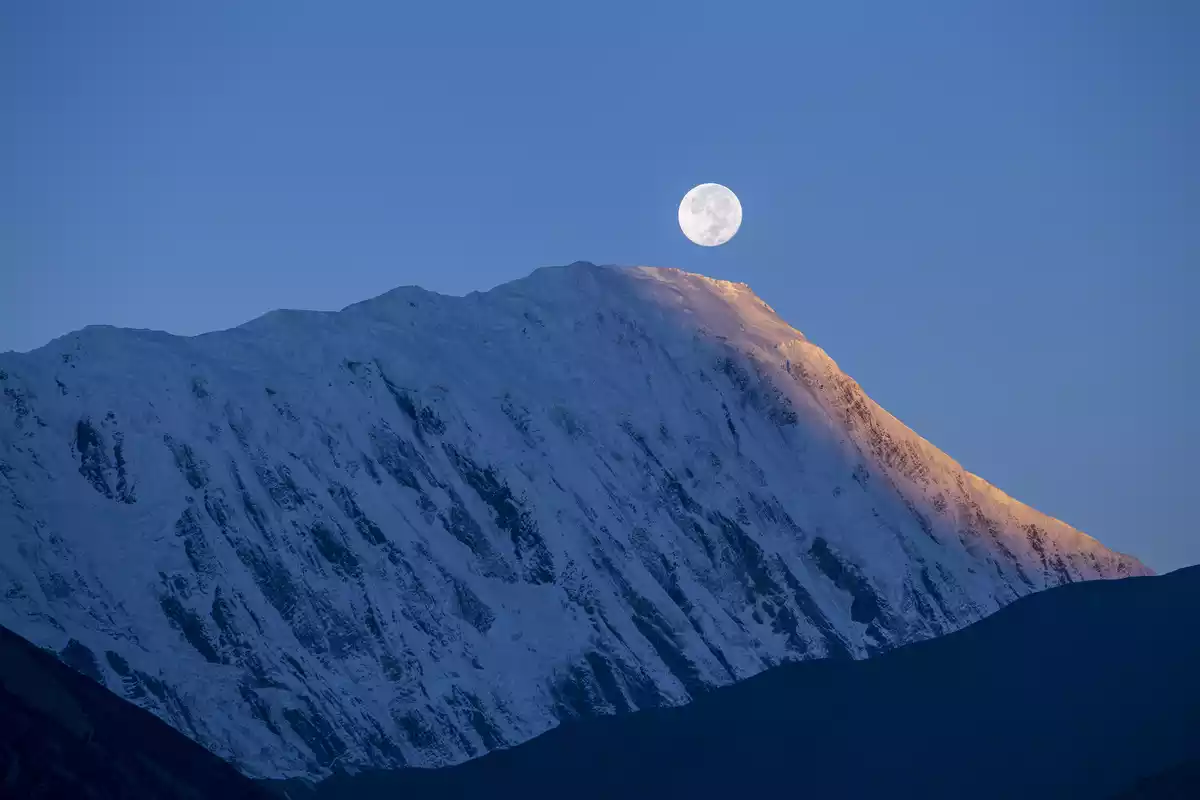 A mountain with snow and the moon illuminated by the sun over it